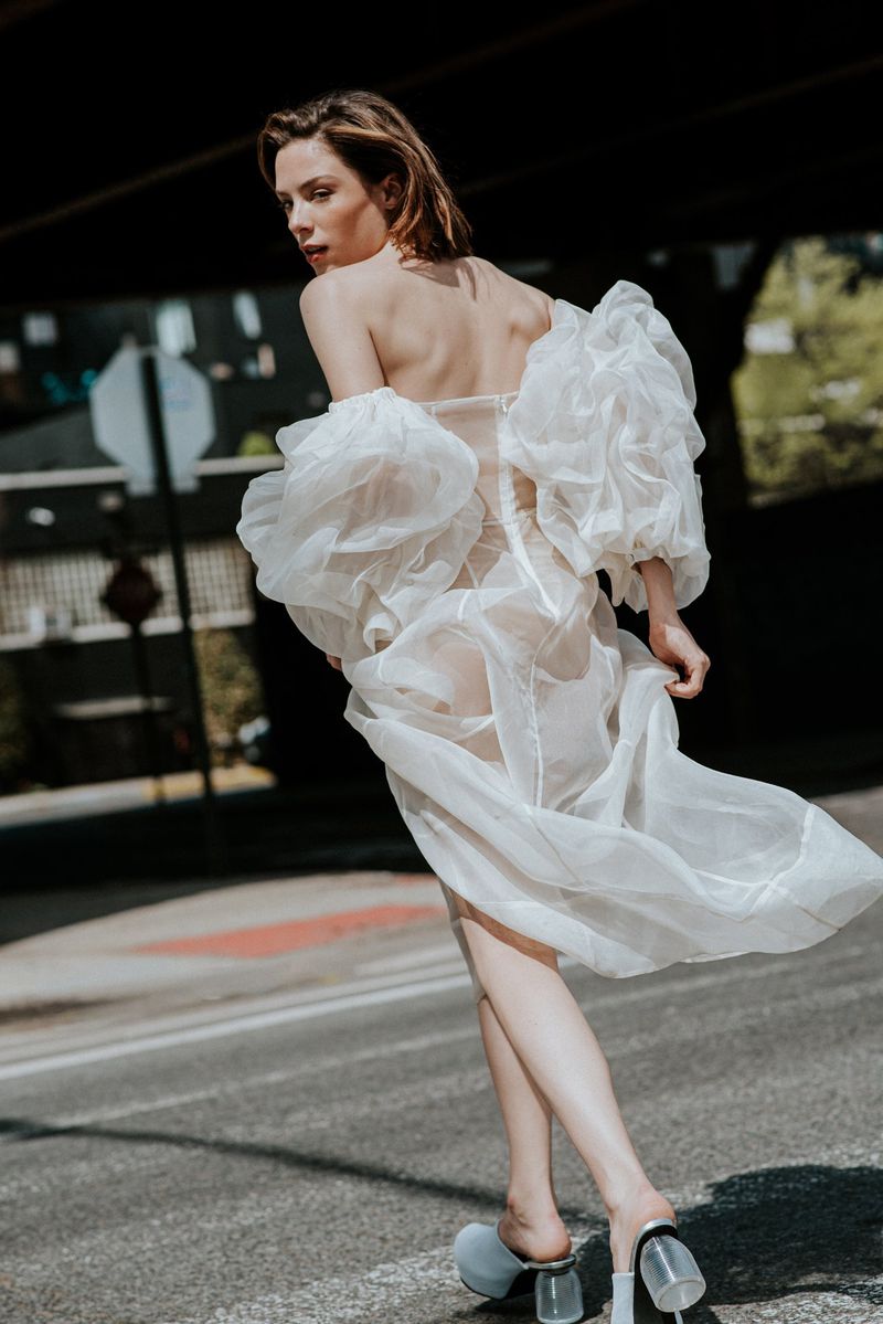 Woman in a sheer white off-the-shoulder dress with voluminous puff sleeves walking on a sunlit city street