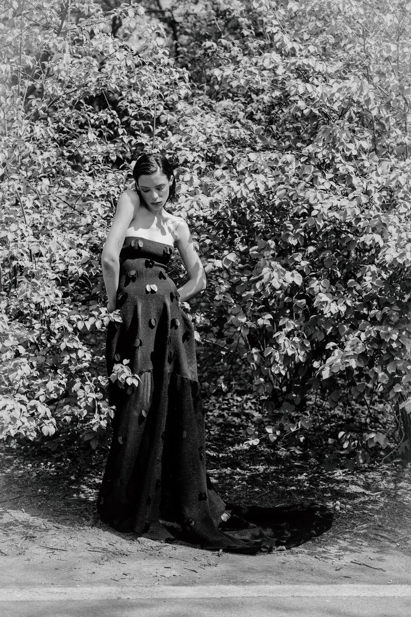 Monochrome fashion portrait of a woman in a strapless black appliqué gown standing against lush leafy bushes outdoors.