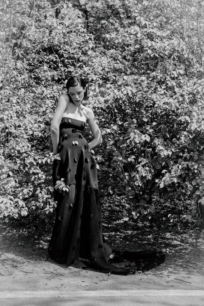 Monochrome fashion portrait of a woman in a strapless black appliqué gown standing against lush leafy bushes outdoors.