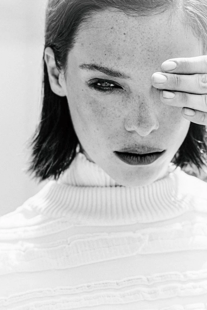 Black and white portrait of a freckled woman in a white turtleneck sweater covering one eye with her hand.