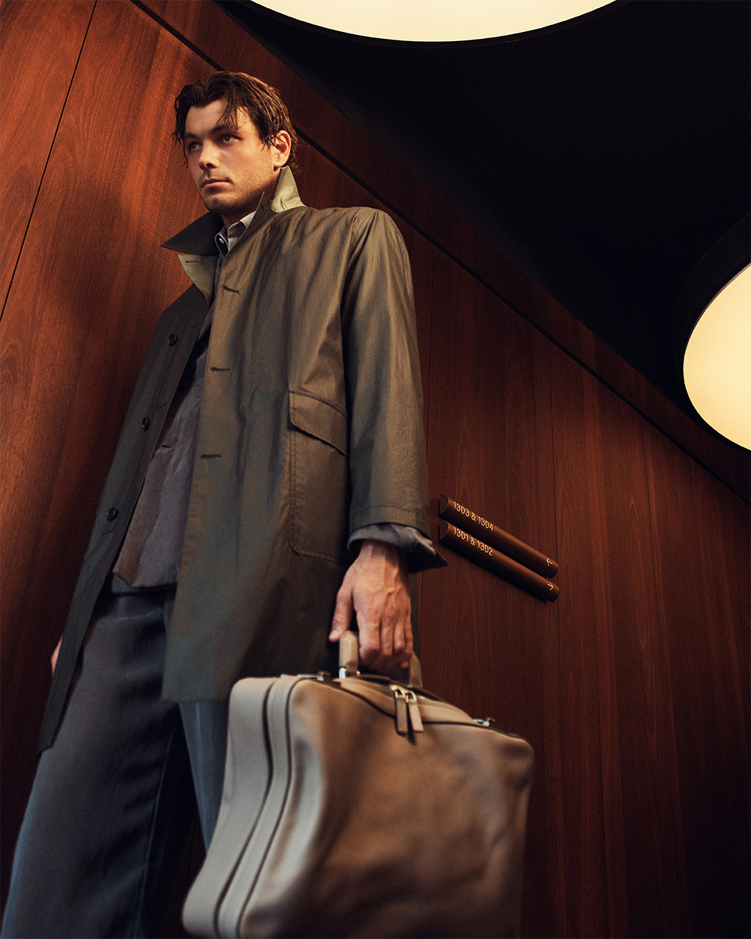 Man in a coat holds a briefcase in an indoor setting with wood paneling and lighting, showcasing a blend of professional style.
