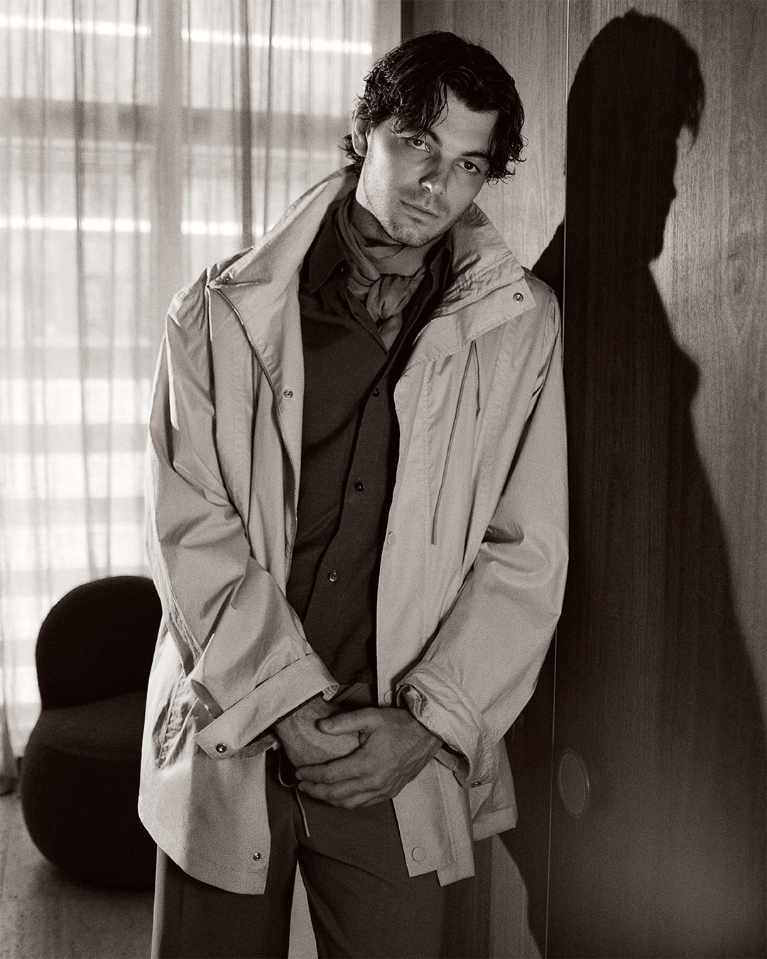 Black and white portrait of a stylish man in an open jacket indoors, posing with a modern chair and natural light from a window.