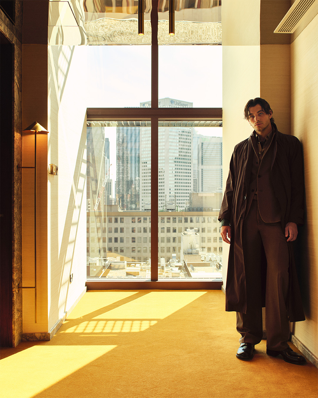 Taylor Fritz wearing a dark brown coat standing in a hotel room with large windows overlooking city buildings