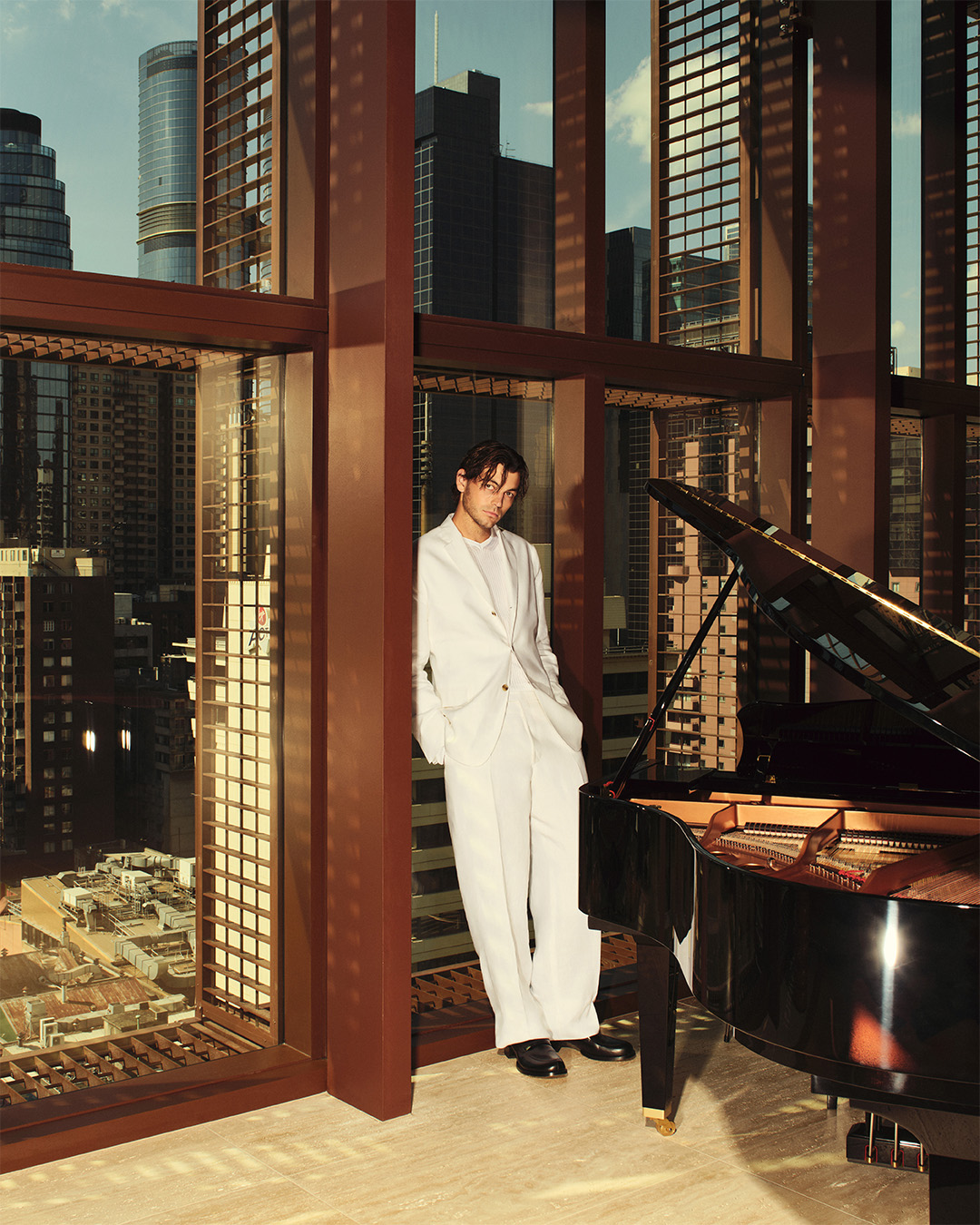Adult male in a white suit poses near a piano in an apartment overlooking a city skyline, bathed in warm light from large windows.