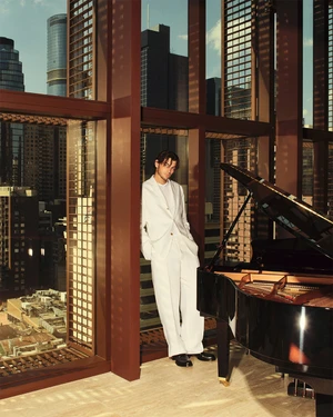 Tennis player Taylor Fritz in a white suit standing near a grand piano in an office overlooking a city skyline