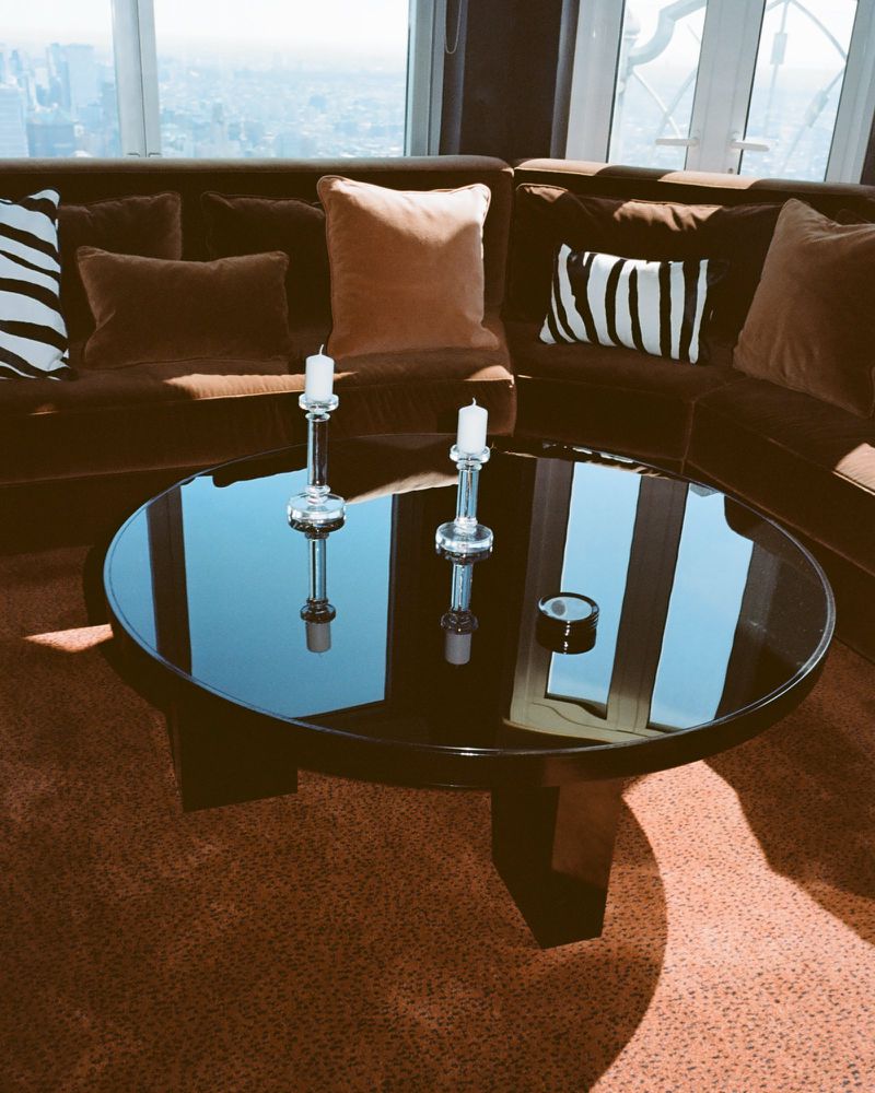 Round black reflective coffee table with glass candle holders set in front of a curved brown velvet sofa with zebra print pillows.