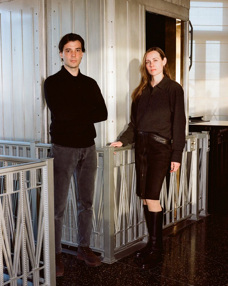 Man and woman in dark attire standing by decorative metal railings in a studio interior.