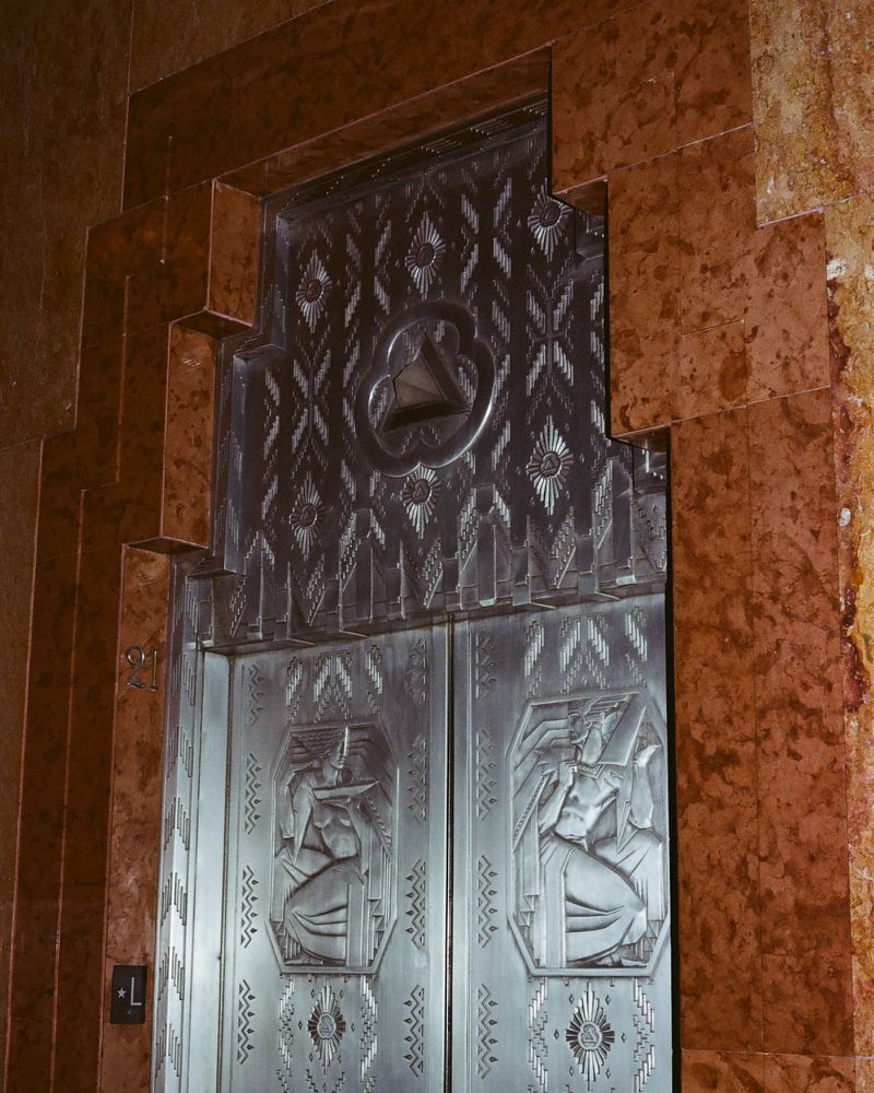 Art Deco embossed silver metal elevator doors set within a stepped reddish brown marble surround.