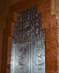 Art Deco embossed silver metal elevator doors set within a stepped reddish brown marble surround.