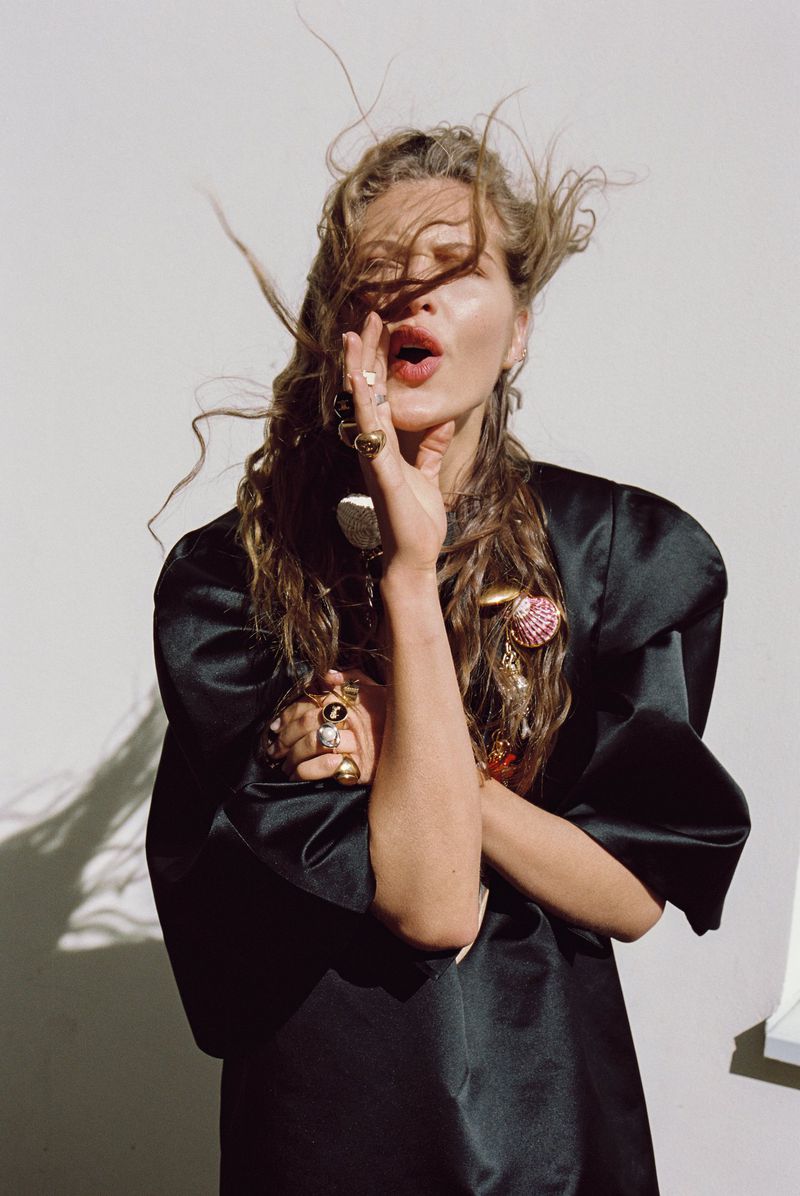 Model with windblown hair wearing a black satin puff-sleeve top and statement gold rings against a sunlit white backdrop.