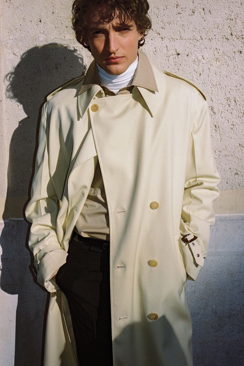 Aliocha Schneider wearing an oversized cream trench coat over a high-collar shirt against a textured stone wall in direct sunlight.