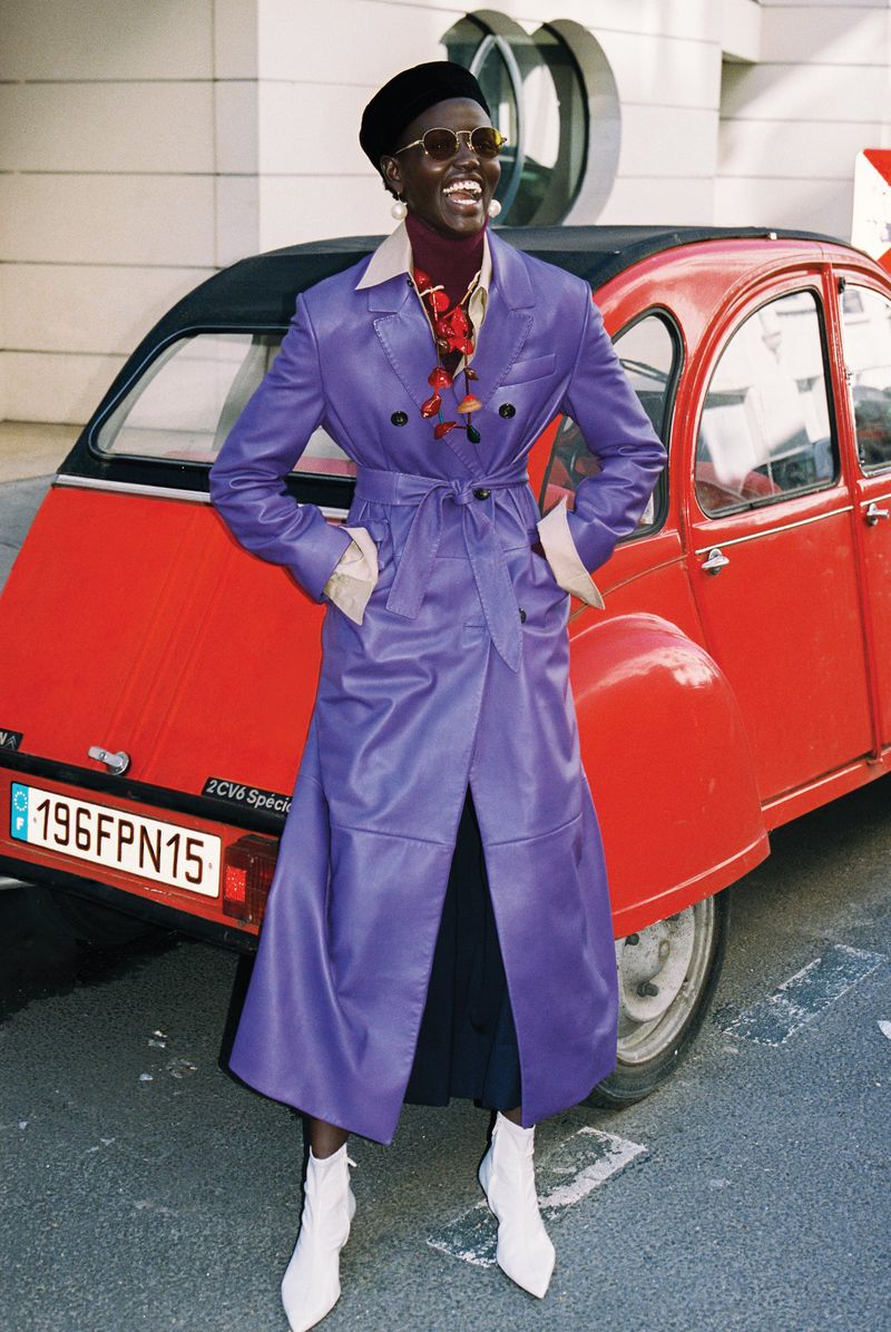 Model Yar Aguer smiling in a purple leather trench coat and black beret in front of a vintage red car.
