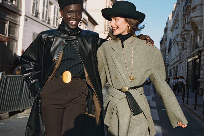Two smiling models walking outdoors wearing black leather coats and hats on a European city street
