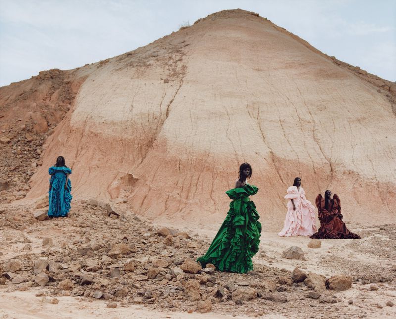 Four models pose in dramatic ruffled ballgowns in green, blue, pink, and brown across a dry, eroded desert mound.