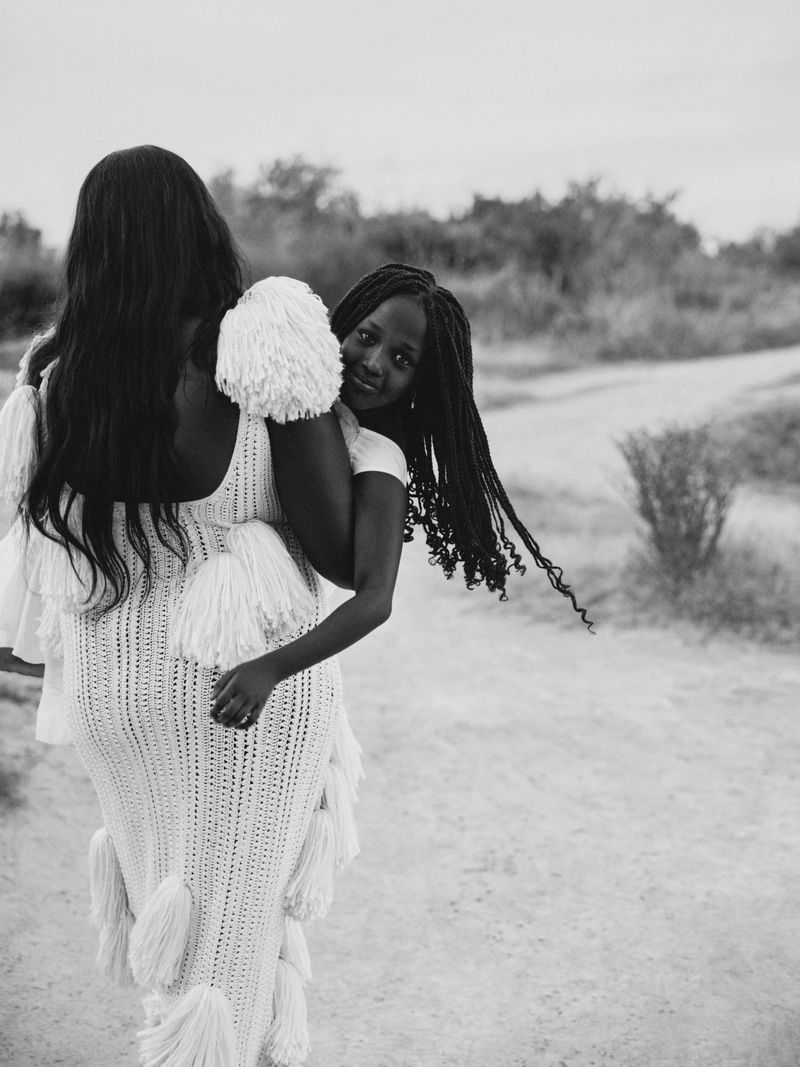 Black and white portrait of a woman carrying a child on her back along an outdoor path for American Vogue.