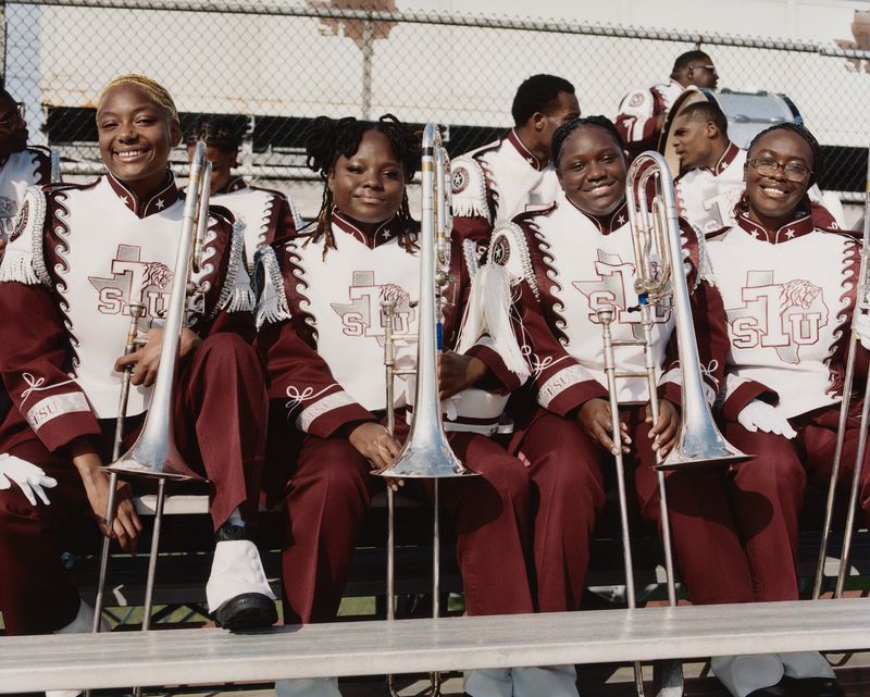 Four young Black women in burgundy and white marching band uniforms holding silver trombones on stadium bleachers.