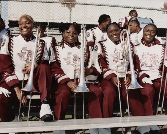 Four young Black women in burgundy and white marching band uniforms holding silver trombones on stadium bleachers.