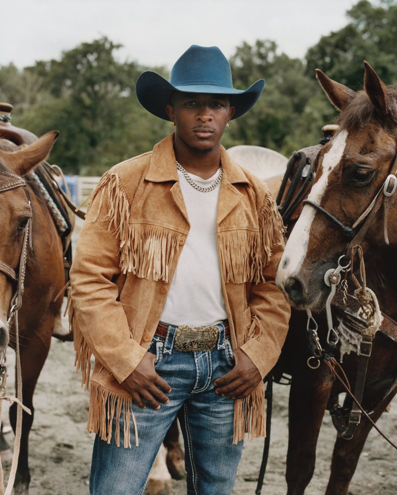 Black man wearing a blue cowboy hat and tan fringe jacket standing outdoors between two saddled horses.