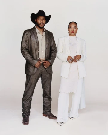 A man in a brown leather suit and black cowboy hat standing beside a woman in a tailored all-white suit against a white backdrop.