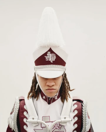 A marching band member with dreadlocks wearing a tall white plumed shako hat and maroon uniform against a plain white background.