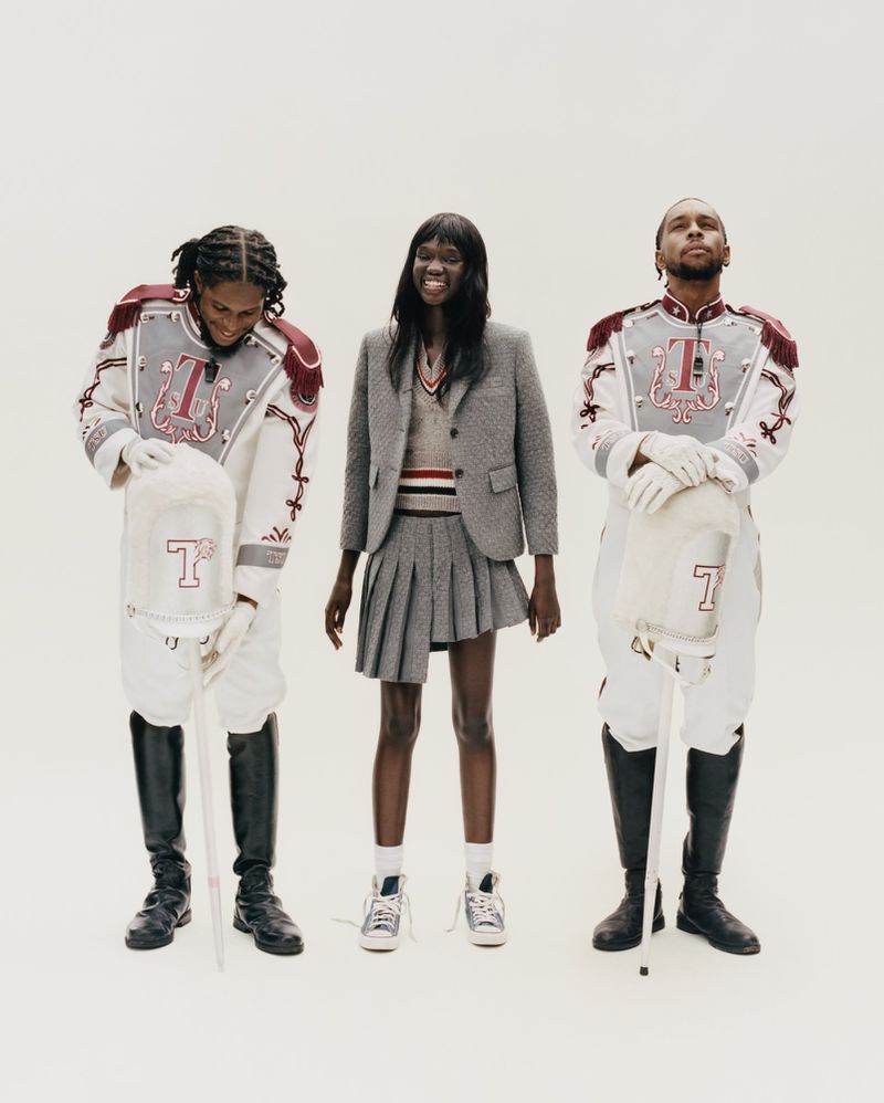 Ruth Deng standing in a grey pleated suit flanked by two men in embroidered marching band uniforms against a white studio background.