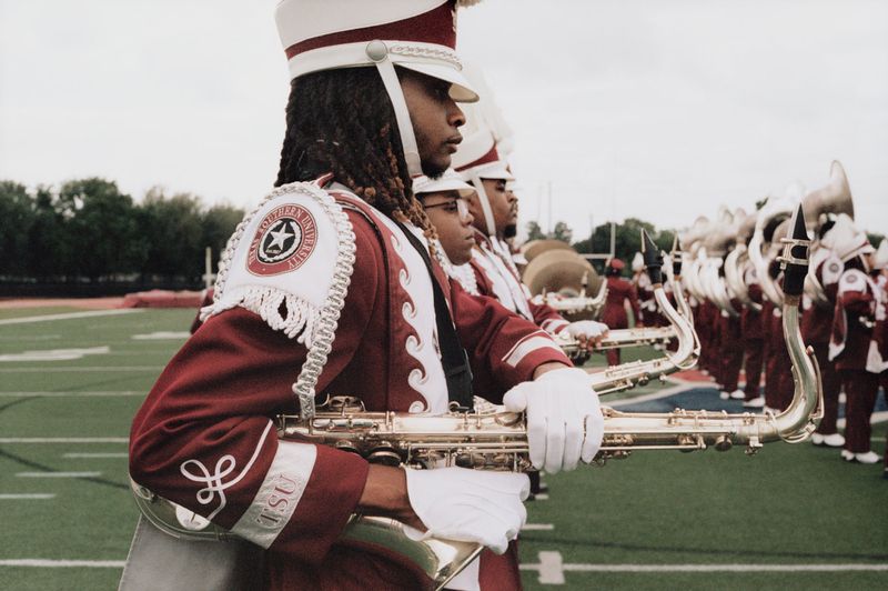 Marching band musicians in maroon and white uniforms holding instruments on a football field for American Vogue