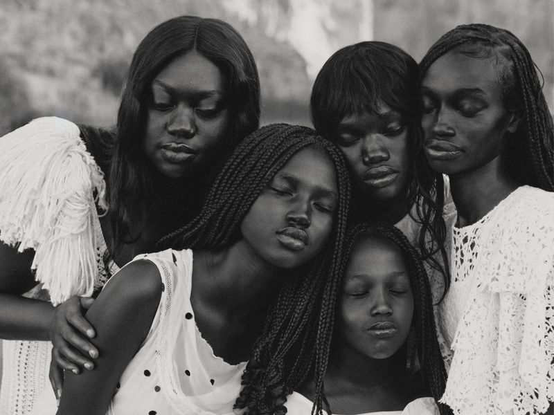 Black and white group portrait of five Black female models in white textured clothing leaning close together with serene expressions.