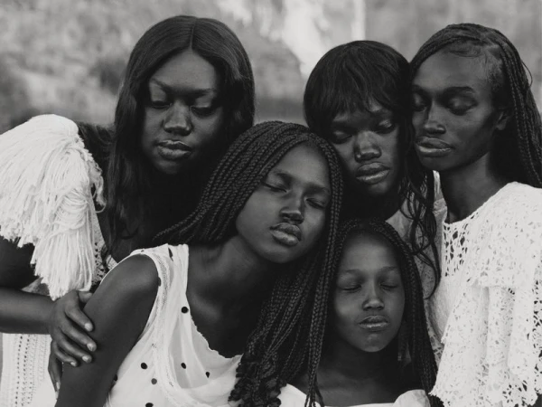 Black and white group portrait of five Black female models in white textured clothing leaning close together with serene expressions.