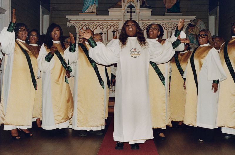 Lead singer and gospel choir members in white and gold robes singing with raised arms before a church altar.