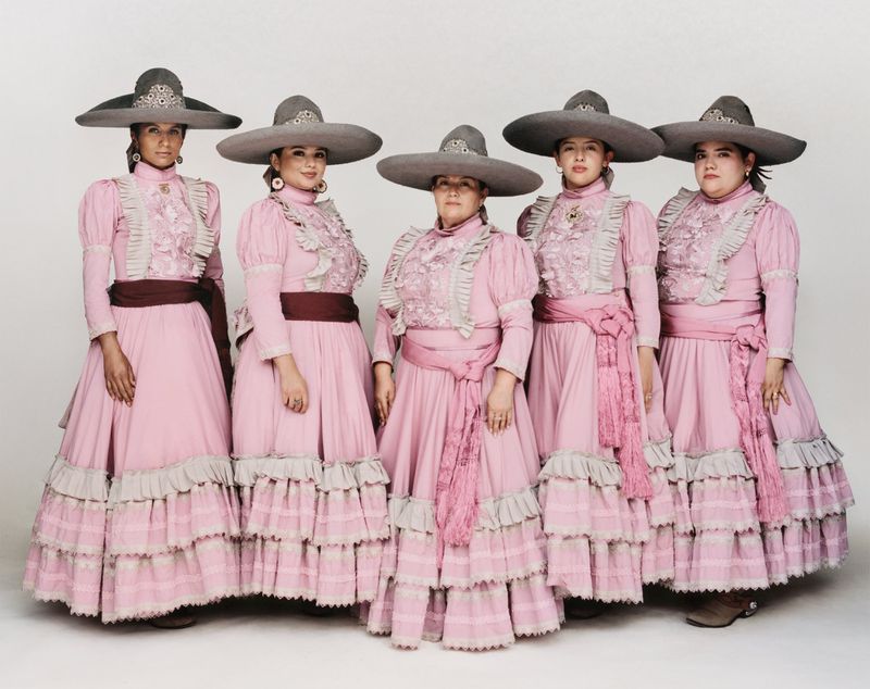 Five women posed in matching pink tiered escaramuza dresses and grey wide-brim charro sombreros against a white studio background.