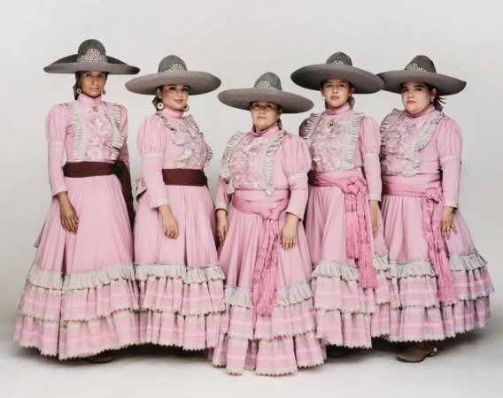 Five women posed in matching pink tiered escaramuza dresses and grey wide-brim charro sombreros against a white studio background.