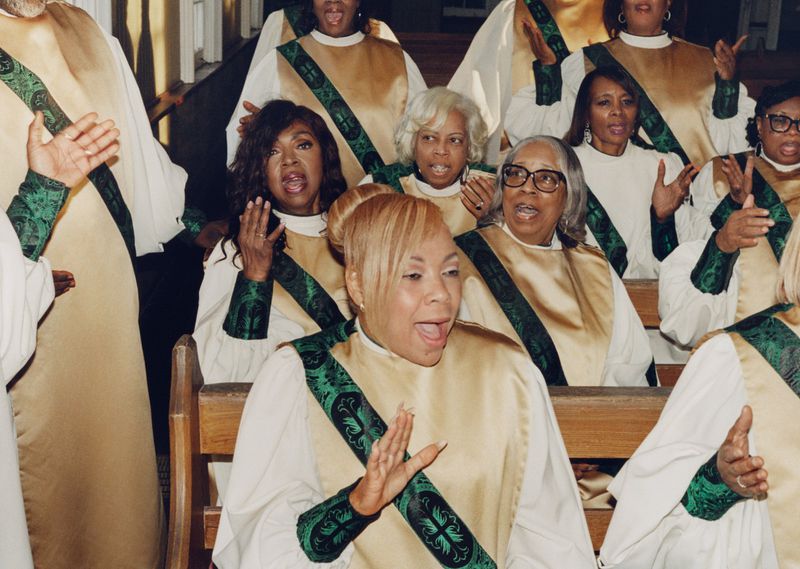 Members of a choir singing in gold and white robes with green patterned stoles while seated in wooden church pews.