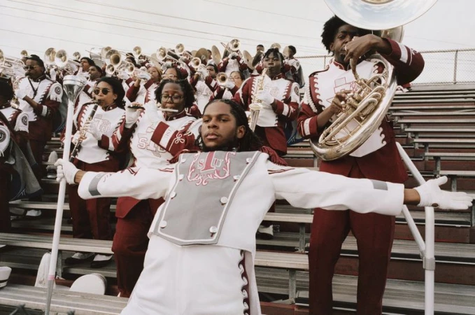 A marching band drum major leaning backward with outstretched arms in uniform on stadium bleachers alongside musicians.