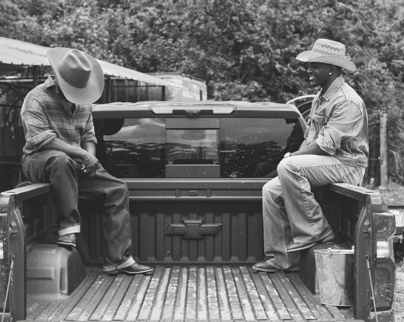 Two Black men wearing cowboy hats and denim sitting on the open bed of a pickup truck in black and white.