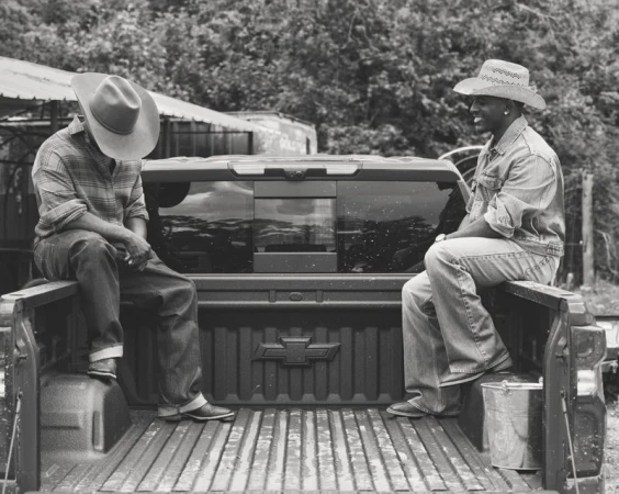 Two Black men wearing cowboy hats and denim sitting on the open bed of a pickup truck in black and white.