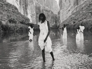 Black women wearing white dresses standing in a shallow river between towering canyon walls in a black and white fashion editorial.