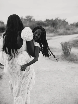 Black and white portrait of a woman carrying a child on her back along an outdoor path for American Vogue.