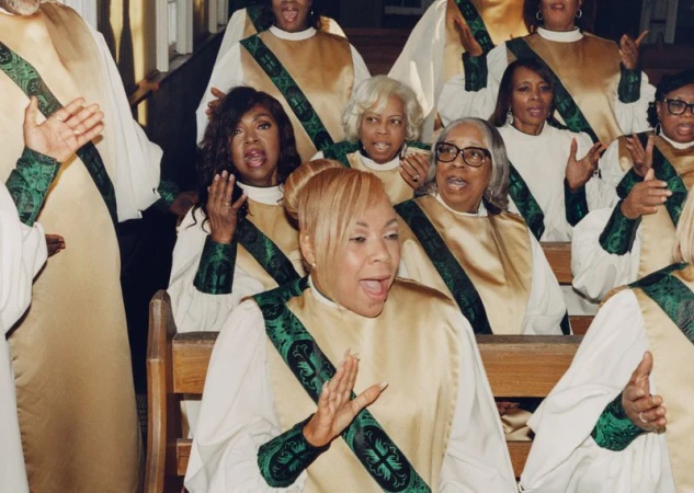 Members of a choir singing in gold and white robes with green patterned stoles while seated in wooden church pews.
