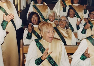 Members of a choir singing in gold and white robes with green patterned stoles while seated in wooden church pews.