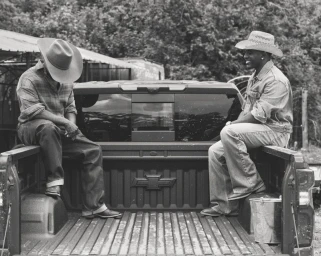 Two Black men wearing cowboy hats and denim sitting on the open bed of a pickup truck in black and white.