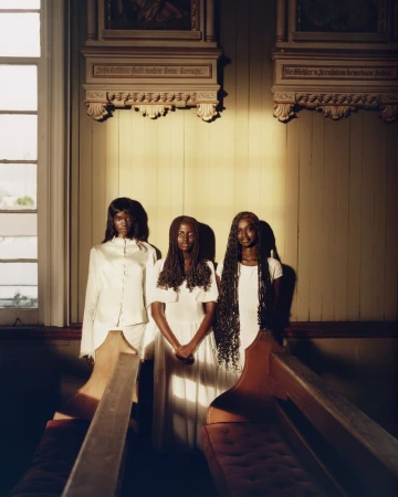 Three Black women in white dresses stand together inside a sunlit wooden church between rows of traditional church pews.