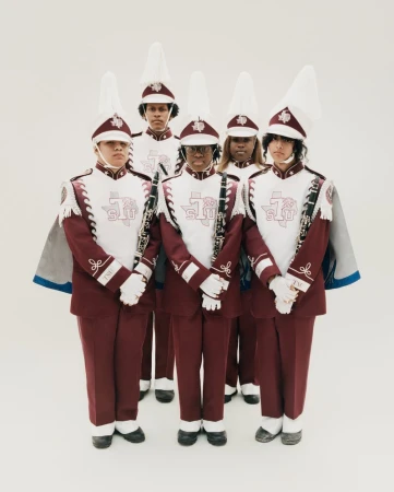 Five marching band members in maroon and white uniforms with tall shako hats stand against a plain white studio background.