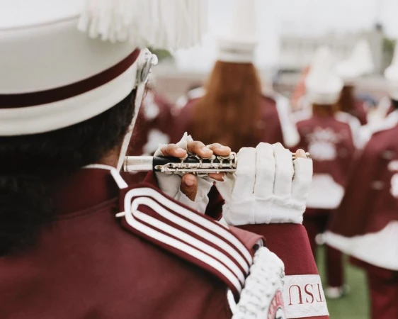 Over-the-shoulder view of a marching band member in a maroon and white uniform playing a silver piccolo outdoors.