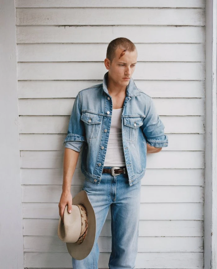 Man in a light wash denim jacket and jeans holding a tan cowboy hat against a white wood paneled wall.