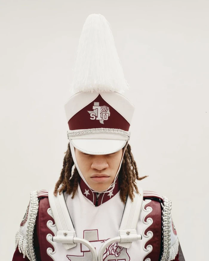 A marching band member with dreadlocks wearing a tall white plumed shako hat and maroon uniform against a plain white background.