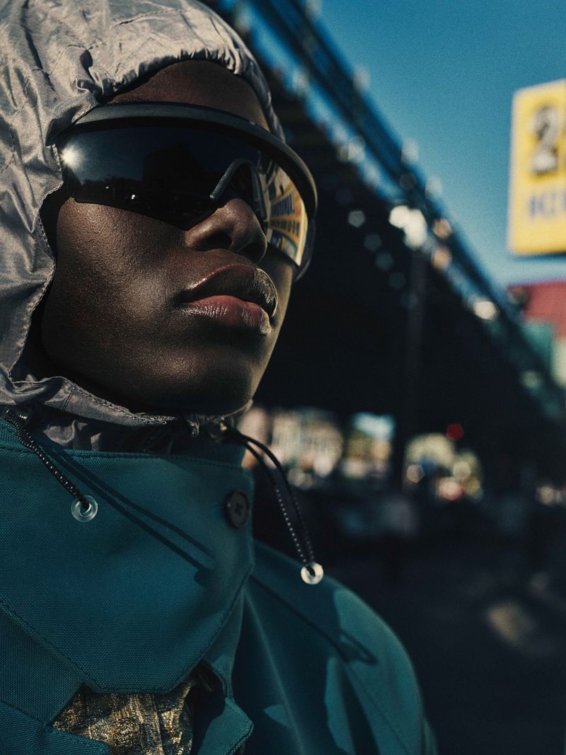 Close-up of a Black model looking up while wearing oversized dark visor sunglasses, a grey hood, and a teal high-collar jacket outdoors.