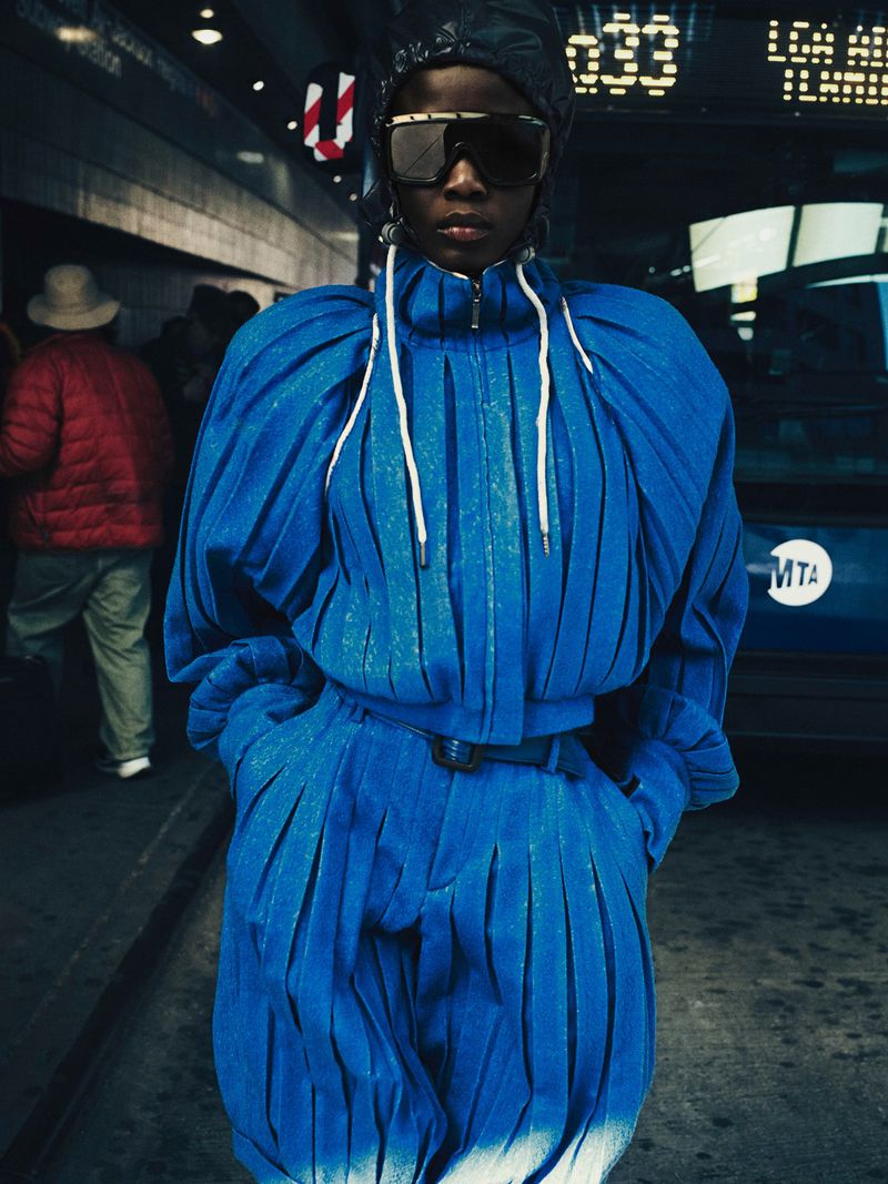 Model Ajah wearing a pleated bright blue tracksuit, black shield sunglasses, and a dark hood in front of a city transit bus.
