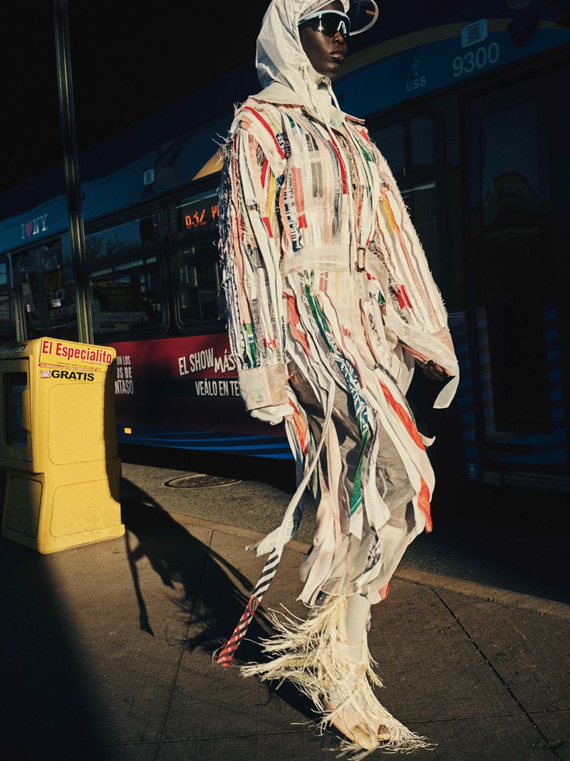 Fashion model in a white fringed coat, hood, and sunglasses standing on a city pavement beside a bus.