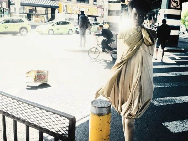 Model Ajah wearing a draped beige dress and metallic gold sash standing on a sunlit city crosswalk in an urban setting.