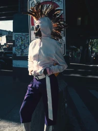Model wearing a feathered headpiece, white hoodie, white sunglasses, and purple cropped trousers on a city street.